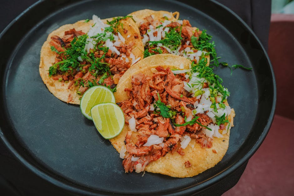 Flat lay of vibrant tacos al pastor with onions, cilantro, and lime on a black plate.